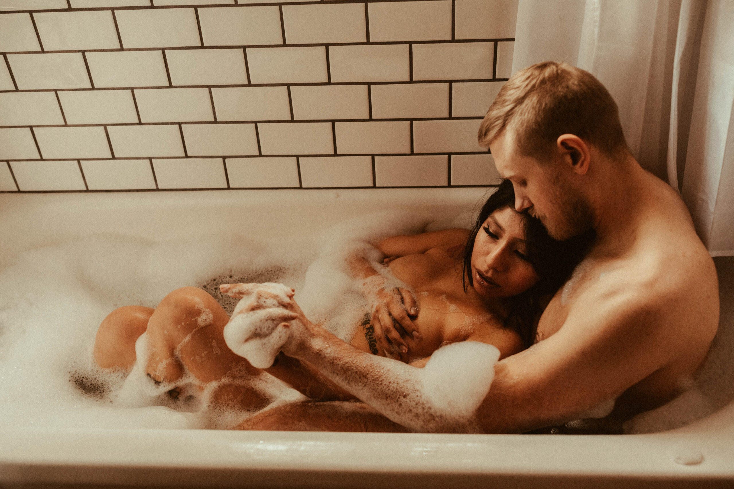couple sitting in bubble bath with arms wrapped each other during steamy couples boudoir photo shoot