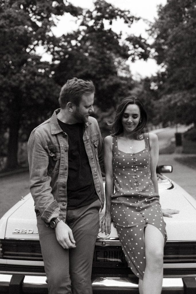 cinematic black and white photo of engaged couple sitting on the back of a vintage car